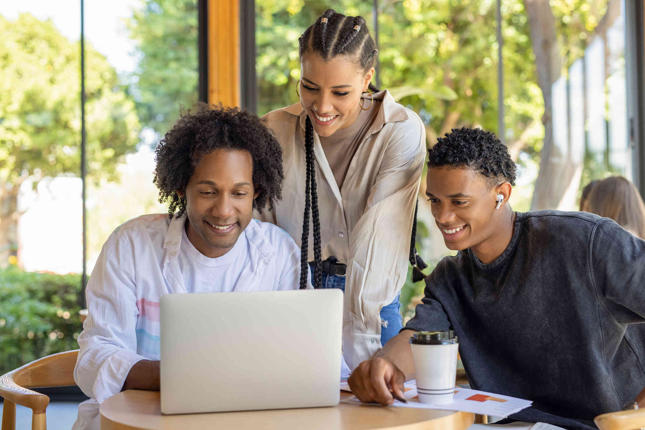 A woman and two men smile as they look down at the screen of a laptop which is sitting on top of a table the two men are sitting at. The woman leans over behind the two men to see.